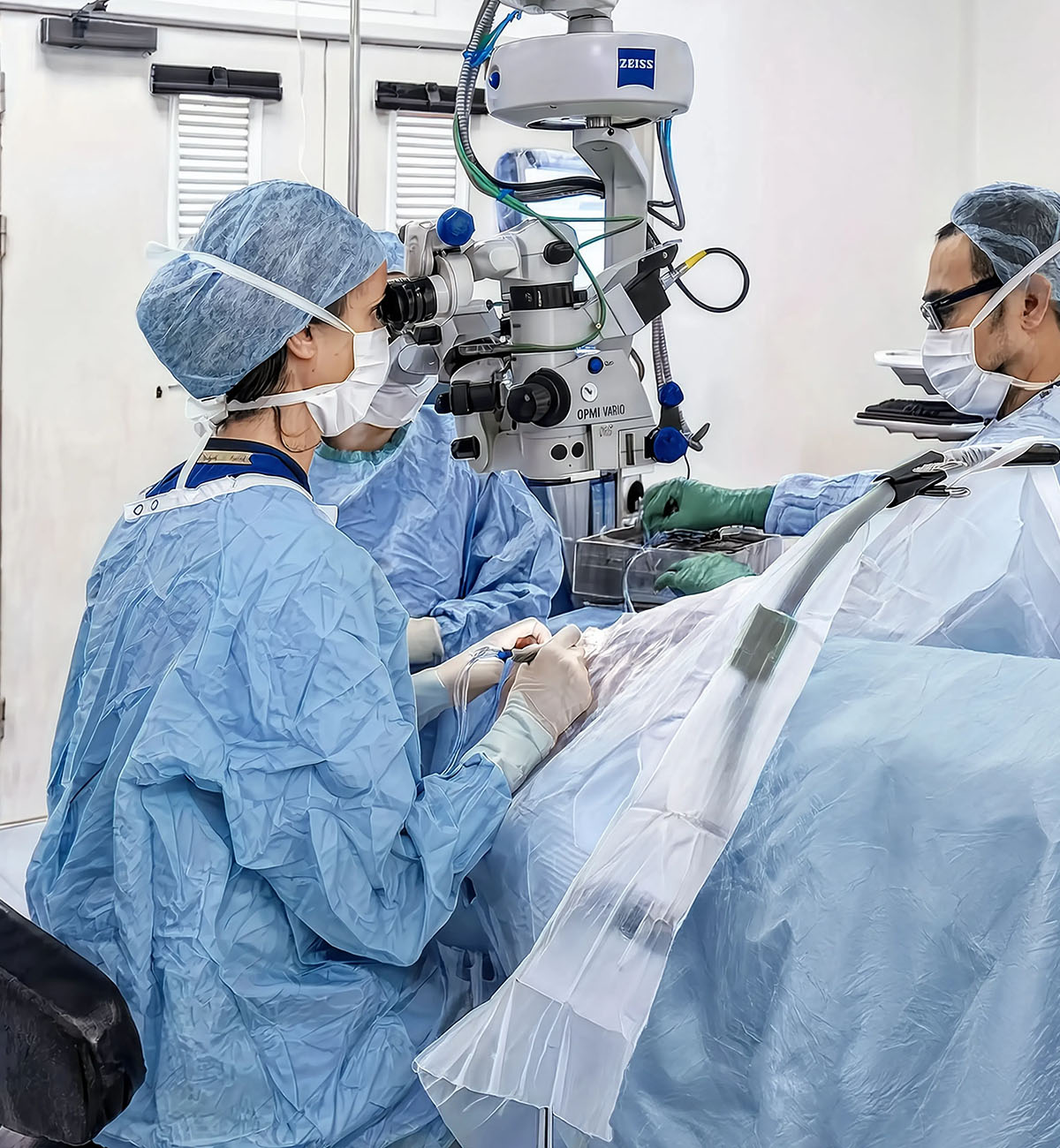 Chirurgo oftalmico esegue un intervento di cataratta guardando attraverso un microscopio operatorio Zeiss OPMI Vario, assistito da un'infermiera con strumenti chirurgici; il paziente è coperto da un telo sterile blu