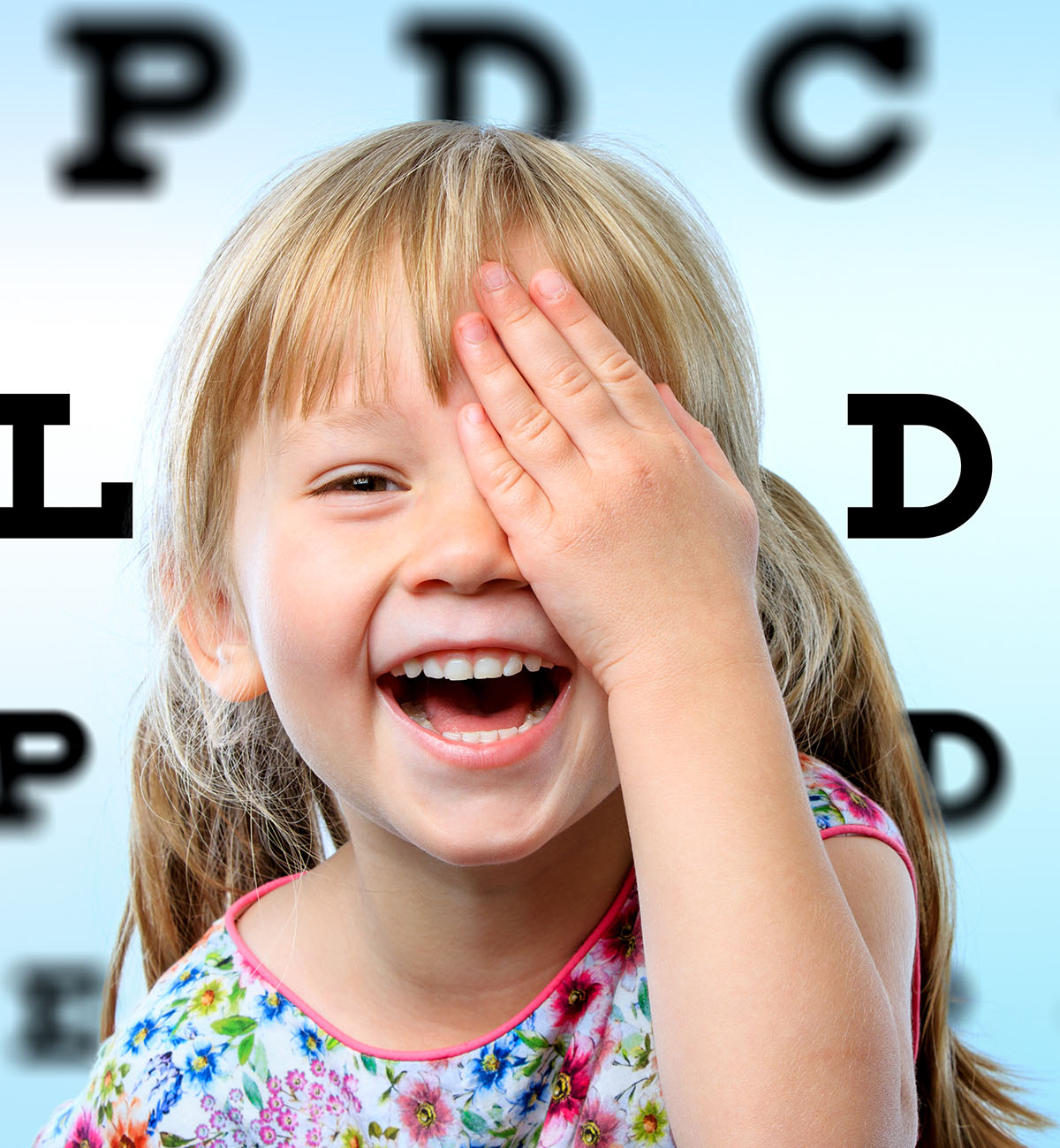 Bambina sorridente si copre un occhio con la mano durante il test della vista, con tabella optometrica sfocata sullo sfondo